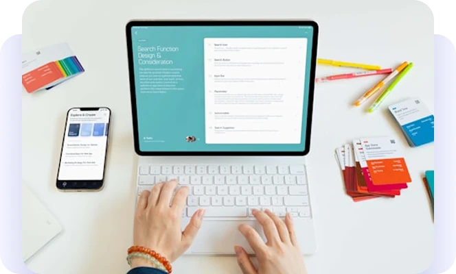 Person using a tablet and keyboard beside a phone, colorful cards, and pencils.