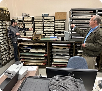 Two employees in an office surrounded by shelves of paper files and materials.
